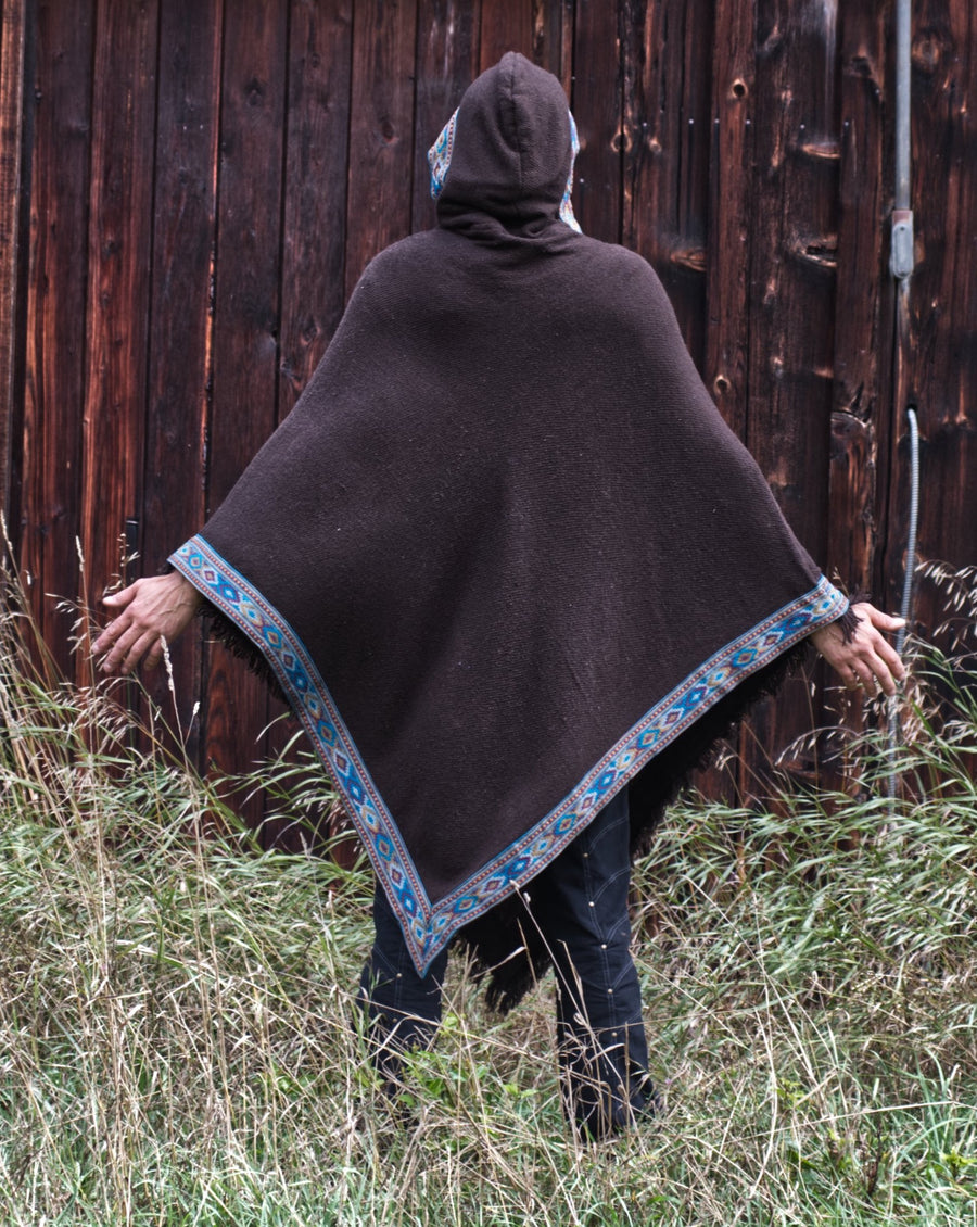 Tall man is standing in front of a barn wall with his back to the camera. His hands are extended to show the width of the poncho and the hood is pulled up over his head.