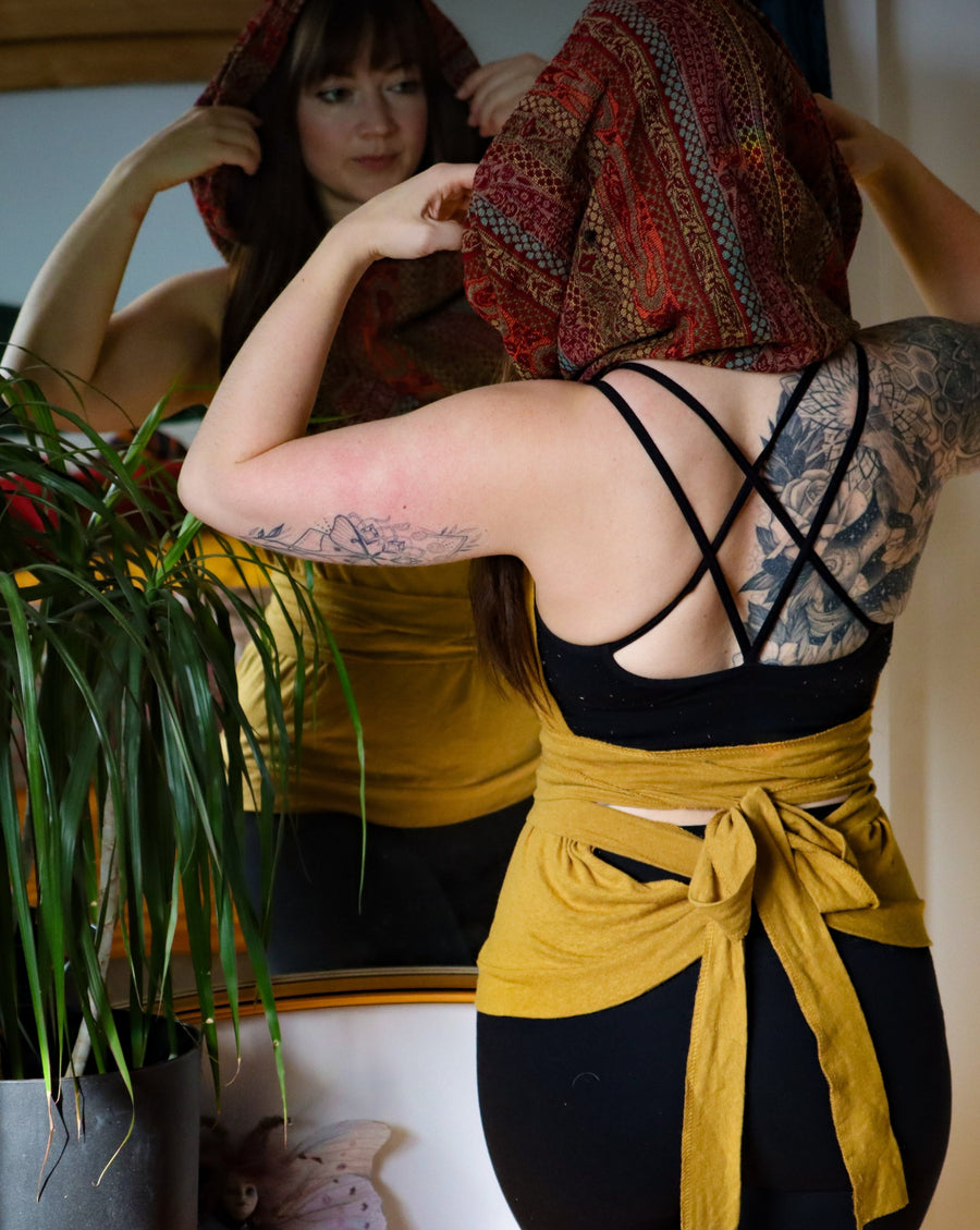 Caucasian girl with back tattoos is standing in front of a mirror wearing a mustard coloured top with a cowl neck. The cowl neck is made of shawl pattern and is complimentary to the mustard. 