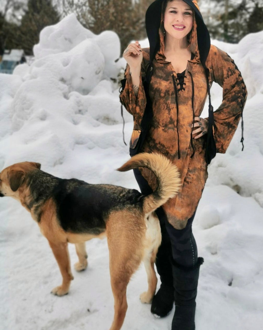 A woman is standing outside surrounded by snow, with one hand on her hip. She's wearing a rusty orange dress, black leggings, Ugg boots and festival fashion wear. Her mix breed german sheppard dog is standing next to her.
