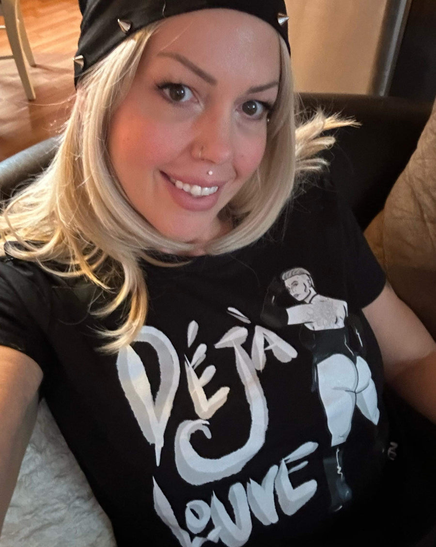 A blond woman with brown eyes is sitting on a couch with a smile that flashes her pearly white teeth. She is waring a black tshirt with the logo of a burlesque performer that says Deja Louve.