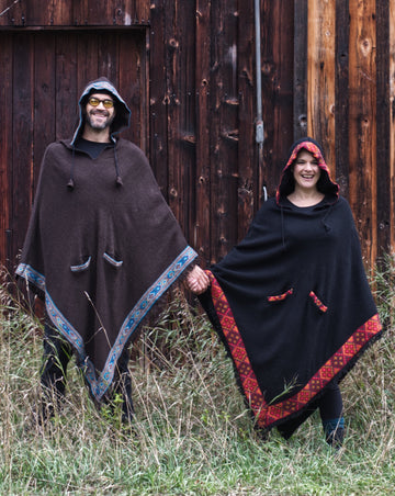 A tall man and a short woman are standing a few feet apart and holding hands while wearing matching ponchos in different colours. Both are smiling.