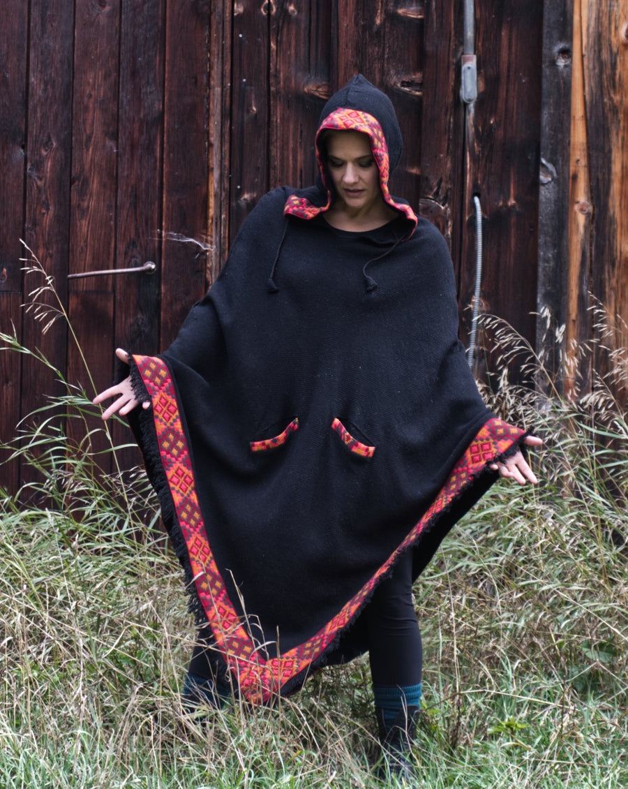 Woman is standing in front of a barn wall while wearing a large, oversized, black poncho. She's holding her hands out to demonstrate the large size of the poncho.