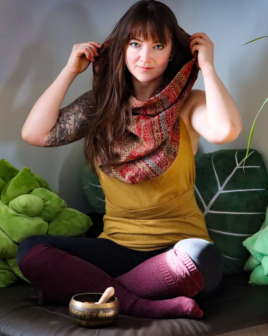 Caucasian girl with brown hair and tattoos on her arms is sitting on a couch surrounded by leaf shaped pillows and is wearing a cotton hemp top with a cowl neck. She's holding up the cowl neck part with her hands.