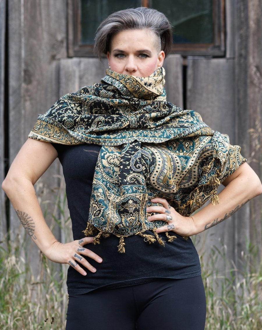 Curvy lady with the sides of her head shaved stands in front of a barn with her hands on her hips while wearing a beautiful scarf wrapped around her neck.