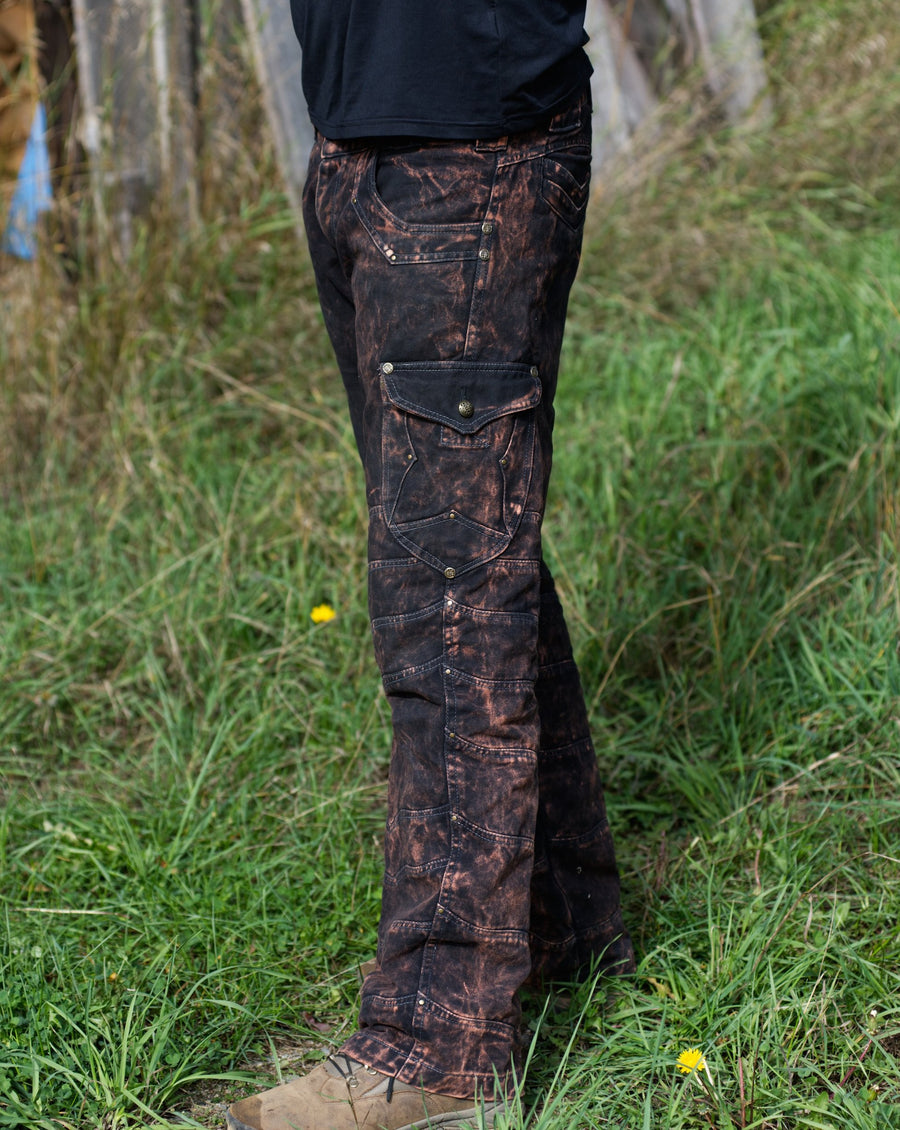 Side view of a man's leg, standing in a field of green grass. The Jeans are an original design, are rust coloured and have many textures and features.