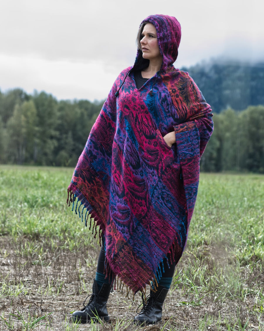 Woman is standing in a field with trees in the background and grey clouds above the trees. She's wearing combat boots and a large, colourful poncho that almost touches the ground. She is looking off to the side.
