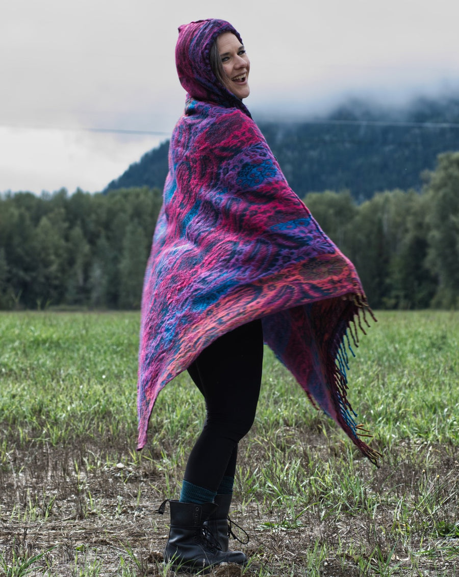 Woman is standing in a field with trees in the background and grey clouds above the tress. She is wearing a large, colourful poncho and is spinning with a smile on her face.