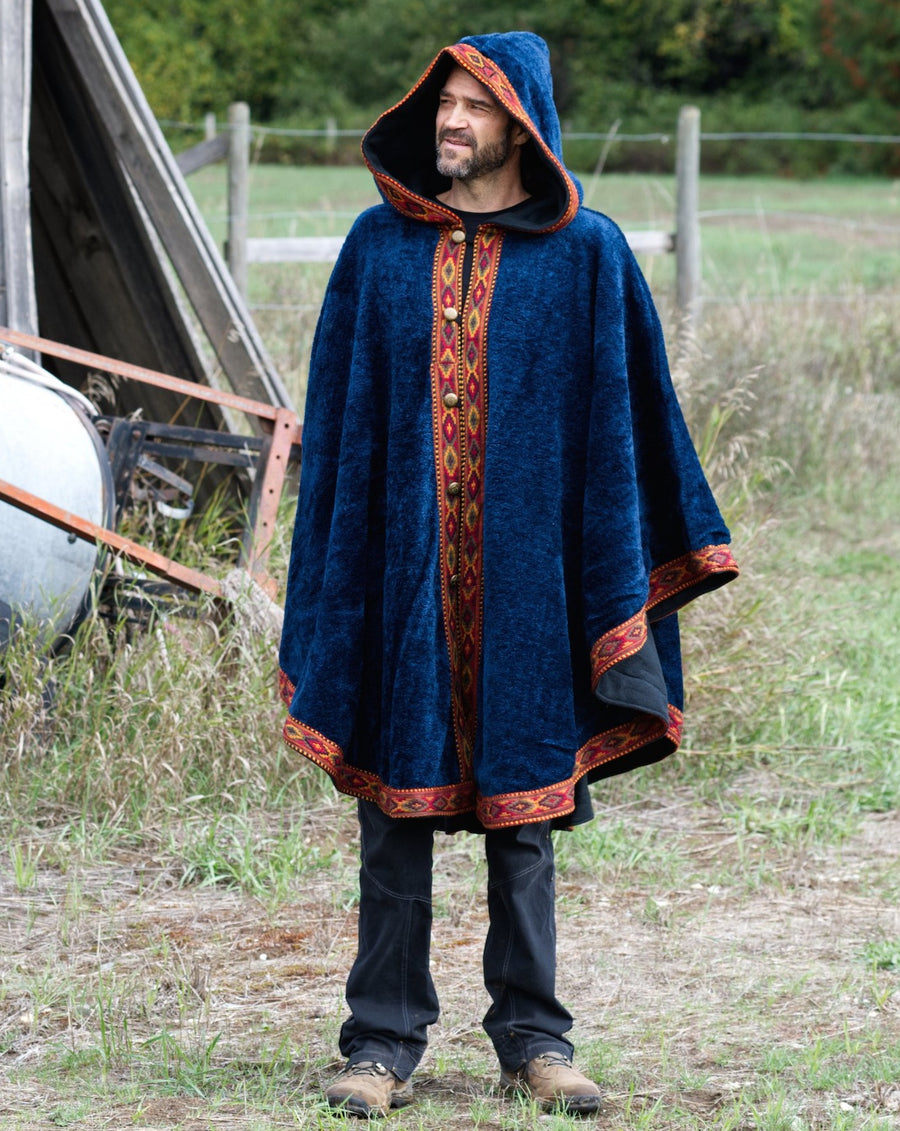 Tall, lean man stands in front of barn with farming equipment in the background while wearing a large, blue, fleece cape, lined with cotton. The cape has a beautiful trim around the edges.