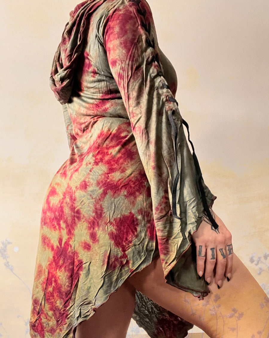 A woman is standing with her back to the camera, her hand hanging down side body, while wearing a long dress with red and green marble thai dye. There is an overlay of wild flowers giving the image a yellow hue and spring vibe.