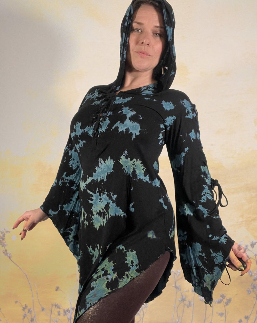 A white woman with short hair is standing with her hands extended out at her hips, in a pretty dancers pose, while wearing a long dress with blue and black marble thai dye. There is an overlay of wild flowers giving the image a yellow hue and spring vibe.